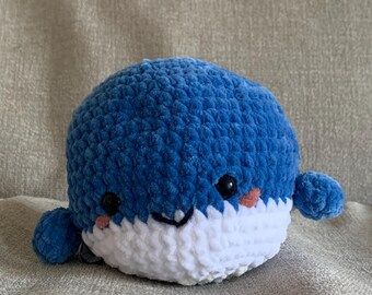 May include: A blue and white crocheted whale plush toy with a smiling face. The whale has small black eyes and pink cheeks.