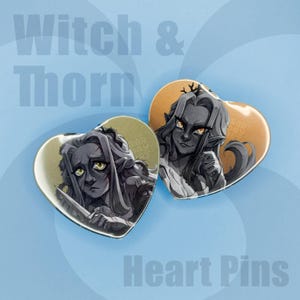 Puede incluir: Dos pines en forma de corazón con ilustraciones de un personaje con piel oscura, pelo largo y ojos amarillos. Un pin muestra al personaje sosteniendo un cuchillo. El texto "Witch & Thorn" y "Heart Pins" son visibles.