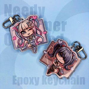 May include: Two epoxy keychains with anime-style characters, each with large eyes and colourful hair. One has pink and blue hair, the other dark hair. Both have heart accents and silver key rings. Text on the image reads "Needy Gamer" and "Epoxy keychain."