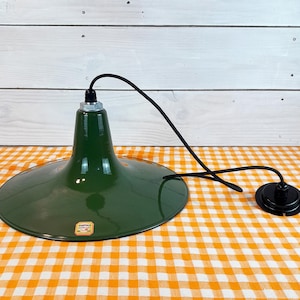 May include: A green metal pendant light fixture with a wide, shallow shade. The shade is a deep forest green, and the fixture has a black cord and a black ceiling mount. The light is sitting on a checkered orange and white tablecloth.