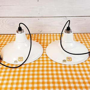 May include: Two white enamel pendant lights with black cords and black metal fixtures hanging from a white and orange checkered tablecloth.