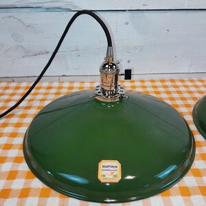 May include: A green metal pendant light fixture with a black cord. The lamp has a shiny, rounded shade and a chrome-colored socket. A label on the shade reads "Benjamin Electric Manufacturing Company". The lamp is on a checkered orange and white surface.