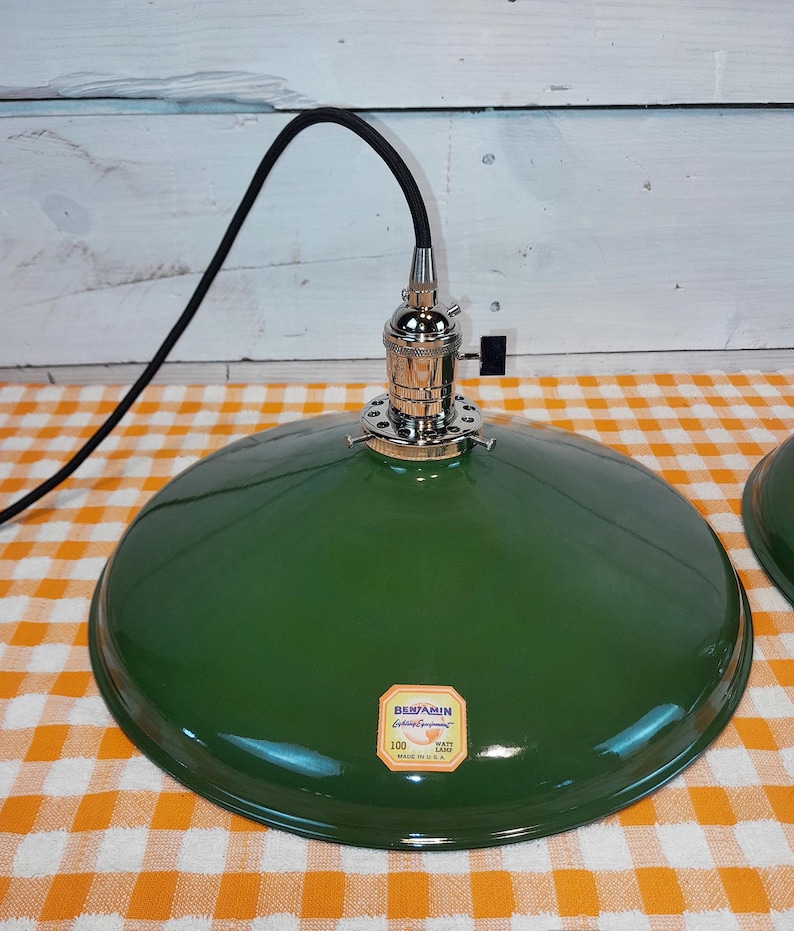 May include: A green metal pendant light fixture with a black cord. The lamp has a shiny, rounded shade and a chrome-colored socket. A label on the shade reads "Benjamin Electric Manufacturing Company". The lamp is on a checkered orange and white surface.