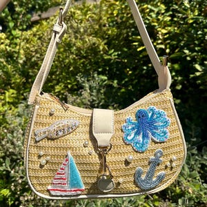 May include: A beige straw handbag with a cream-colored strap and accents. The bag features beaded embellishments of a fish, sailboat, octopus, and anchor, along with scattered faux pearls. The bag has a zipper closure and a cream-colored flap.