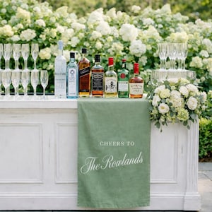 May include: A white bar with various liquor bottles and champagne flutes. A green linen cloth with the text "Cheers to The Rowlands" is draped over the front. White flowers and greenery are arranged on the bar.