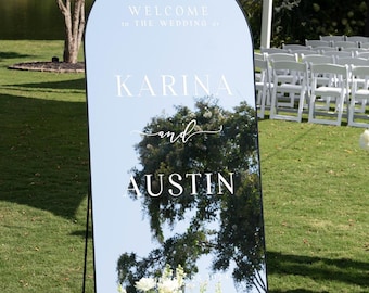 May include: A black arched mirror with a white sign that reads "Welcome to the wedding of Karina and Austin". The mirror is standing on a grassy lawn with white flowers in front of it.