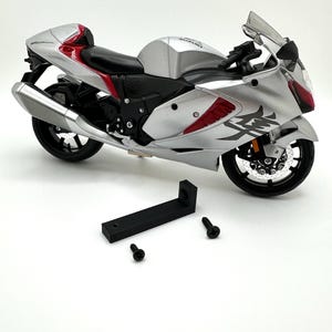 Könnte beinhalten: Ein silbernes und rotes Suzuki Hayabusa Motorradmodell mit schwarzen Rädern und einem schwarzen Kunststoffständer. Das Motorrad hat einen schwarzen Sitz und einen schwarzen Auspuff. Das Motorrad steht auf einer weißen Oberfläche.