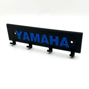 Puede incluir: Porta llaves negro con la palabra "YAMAHA" en letras azules. El porta llaves tiene cuatro ganchos.