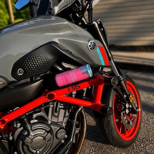 Puede incluir: Una motocicleta Yamaha MT-07 gris con una jaula de protección de color rojo brillante con el texto FMS impreso. La moto tiene ruedas rojas, un bloque de motor negro y una pequeña lata de bebida rosa sobre el marco rojo. El depósito es gris.