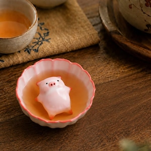 Puede incluir: Una pequeña figura de cerdo de cerámica rosa claro descansa dentro de una taza de té festoneada, blanca y rosa, llena de líquido color ámbar. El cerdo tiene un acabado liso y brillante con sutiles tonos anaranjados en orejas y patas.