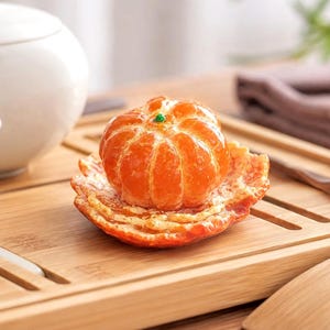 Puede incluir: Una escultura de cerámica realista de una mandarina pelada que descansa sobre una base de cáscara de naranja texturizada. La fruta presenta una superficie naranja brillante y vibrante con segmentos distintos y un pequeño tallo verde arriba.