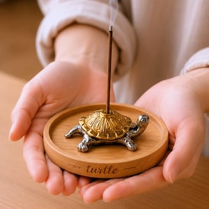 Könnte beinhalten: Ein runder Räucherstäbchenhalter aus Holz mit einer metallischen Schildkrötenfigur, die einen goldfarbenen Panzer und einen silberfarbenen Körper hat. Das Wort turtle ist in den vorderen Rand des Holzsockels eingraviert, der ein Räucherstäbchen hält.
