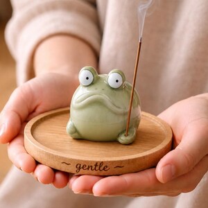 Könnte beinhalten: Ein hellgrüner Keramik-Frosch-Räucherstäbchenhalter mit großen weißen Augen, der auf einem kleinen Holztablett sitzt. Auf dem Tablett steht das Wort "gentle". Ein Räucherstäbchen ist angezündet, Rauch steigt auf.
