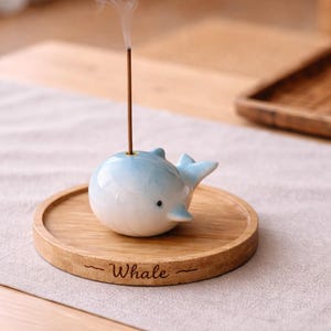 以下が含まれることがあります： 青と白のセラミック製のクジラ型お香立て。お香が焚かれて煙が出ています。お香立ては、"Whale"の文字が刻まれた丸い木製のトレイの上に置かれています。
