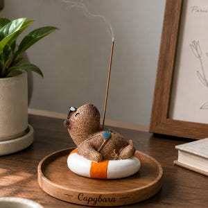 Può includere: Una statuetta di capibara marrone che riposa in un salvagente bianco e arancione su un vassoio rotondo di legno. Il capibara indossa occhiali blu e ha un bastoncino di incenso inserito sulla schiena, che emette una sottile scia di fumo. La parola Capybara è incisa sul vassoio.