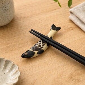 Pode incluir: Um par de pauzinhos pretos mate sobre um suporte de cerâmica em forma de peixe. O suporte apresenta um corpo de cor creme com marcas pintadas a preto e um padrão de grelha no dorso, com um acabamento de verniz brilhante.