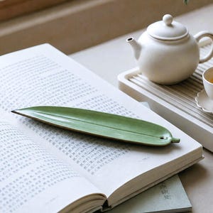 Puede incluir: Una bandeja de cerámica verde, larga y delgada, con forma de hoja, punta afilada y un pequeño orificio en la base. La bandeja descansa sobre un libro abierto con texto impreso. Al fondo, una tetera blanca se sitúa sobre una bandeja de listones.