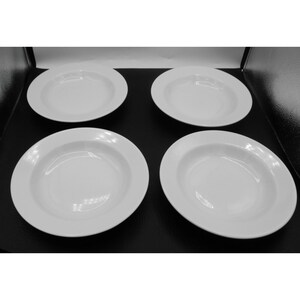 May include: Four white, round, shallow soup bowls with a wide rim are arranged on a black surface. The bowls are evenly spaced and appear to be made of ceramic or porcelain.