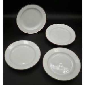 May include: Four white ceramic plates with a brown rim. One plate is displayed on a metal stand. The plates are round and have a simple, classic design. The plates are suitable for serving meals.