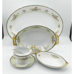 May include: A vintage porcelain serving set featuring an oval platter, two serving dishes, and a gravy boat with a saucer. The set has a white base with a pale yellow border, gold trim, and floral accents in pink, yellow, and green.