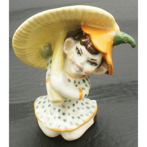 May include: A ceramic figurine of a small child wearing a mushroom hat. The figurine is white with blue polka dots on the dress and orange accents. The mushroom is yellow and orange with a green stem.