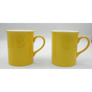 May include: Two yellow ceramic mugs with white interiors and curved handles. The mugs have a smooth, glossy finish and are set against a plain white background. The mugs are identical in shape and size.