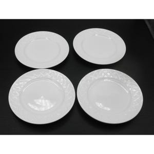 May include: Four white ceramic plates with a textured, woven-like pattern around the rim. The plates are round with a smooth, plain center. The plates are arranged on a dark surface.