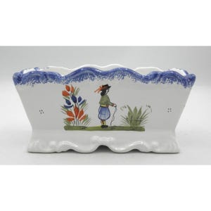 May include: A rectangular white ceramic planter with a scalloped rim featuring a blue sponge-painted edge. The front displays a hand-painted illustration of a figure in a green jacket and blue striped trousers standing between colorful plants.