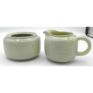 May include: A light green ceramic sugar bowl with a lid and a matching creamer. Both pieces have a ribbed design. The creamer has a handle and a spout. The set is likely for serving coffee or tea.