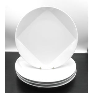May include: A stack of three white ceramic plates featuring a distinct geometric design. Each plate has a smooth, glossy finish with a square-shaped center panel that transitions into rounded edges, creating a clean and uniform appearance.