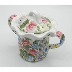 May include: A white ceramic floral-patterned sugar bowl with a lid and handle. The bowl and lid are adorned with pink, red, blue, and purple flowers and green leaves. The handle is curved and the bowl has a matching handle.