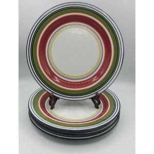 May include: A stack of white ceramic plates with colorful striped rims. The plates feature concentric rings of red, green, yellow, and blue. The plates are stacked on a wooden plate stand, with a white background.