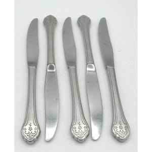 May include: A set of five silver-toned metal dinner knives arranged vertically. Each knife features a smooth, curved blade and a handle decorated with an embossed floral pattern at the base and vertical lines extending up the center of the handle.