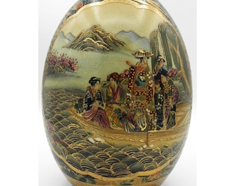 May include: An oval-shaped decorative egg with a detailed Japanese scene. The egg features a boat with figures, mountains, and water, all painted in vibrant colors with gold accents. The top and bottom have intricate patterns.