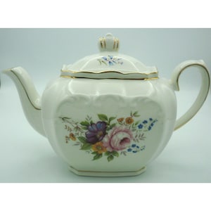 May include: White porcelain teapot with a floral design featuring purple, pink, and yellow flowers. The teapot has a gold trim, a curved handle, and a lid with a gold crown-like finial. The spout is curved and elegant.