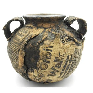 May include: A handcrafted ceramic vase with two handles, featuring a unique design of layered newspaper clippings. The vase has a textured, earthy appearance with dark brown and tan tones. The text on the newspaper is partially visible.