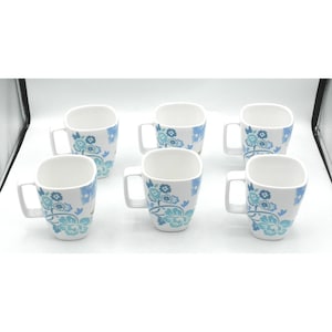 May include: A set of six white ceramic mugs, each featuring a square-shaped body and a handle. The exterior of every mug is decorated with a repeating pattern of blue and light blue floral motifs, including stylized blossoms and leafy vines.