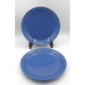 May include: Two round, solid blue ceramic plates featuring a smooth, glossy finish and a decorative embossed beaded pattern along the outer rim. One plate is positioned upright on a small dark brown stand, while the second plate lies flat.