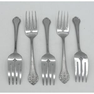 May include: A set of five silver-toned metal forks arranged in a row. Each fork features a three-tined head and a handle with a decorative floral pattern at the base. The forks are uniform in size and shape, displaying a polished metallic finish.