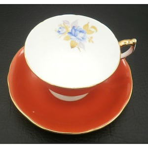 May include: A vintage teacup and saucer set. The teacup is white with a blue floral design and gold trim on the handle. The saucer and the exterior of the teacup are a vibrant orange-red color, with gold trim. The set is likely made of porcelain.