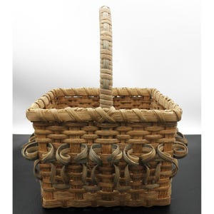 May include: A square, woven basket with a handle. The basket is made of light brown and tan woven material. The handle is vertical and the basket has decorative loops around the sides.