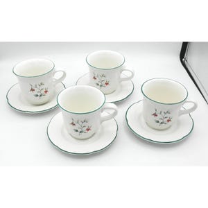 May include: Four white teacups and saucers with a green trim. Each cup and saucer features a floral design with red berries and green leaves. The cups have a handle and are placed on a white surface.
