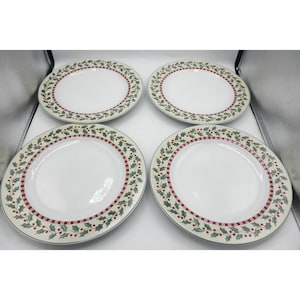 May include: A set of four round white dinner plates, each featuring a decorative border pattern of green holly leaves and small red berries, accented by a thin red and white checkered line encircling the inner rim of the plate surface.