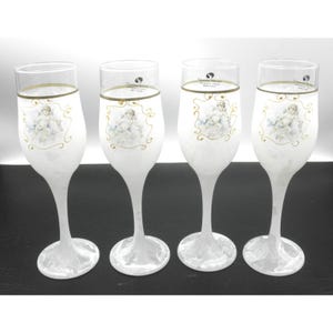 May include: Four champagne flutes with a frosted white finish. Each glass features a gold rim and a decorative image of an angel. The glasses have a slender stem and a wide, round base. The glasses are arranged in a row.