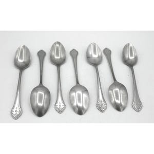 May include: A set of seven silver-toned metal spoons arranged in a row. Four spoons feature a decorative floral engraving at the base of the handle, while three spoons have a plain, smooth handle design with a small pointed tip at the end.