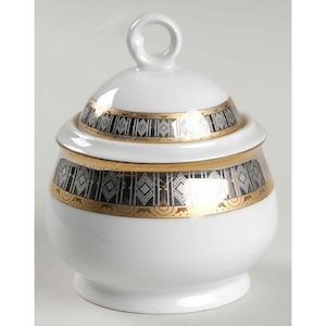 May include: A white ceramic sugar bowl with a lid and a loop handle. The bowl and lid are decorated with a gold and silver geometric pattern. The bowl has a round base and a slightly flared rim.