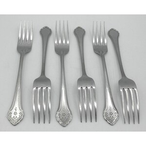 May include: A set of six silver-toned metal forks arranged in two rows. Three forks feature a decorative floral engraving on the handle base, while the other three have plain, smooth handles with a rounded, scalloped tip design.