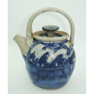 May include: A ceramic teapot with a blue and white wave pattern. The teapot has a curved spout, a rounded body, and a handle. The lid is topped with a small knob. The overall design is rustic and handmade.