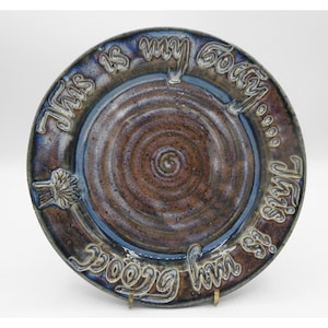May include: A handmade ceramic plate with a spiral design in the center. The plate has a brown and blue glaze with the words "This is my body" inscribed around the rim. The plate is supported by a metal stand.