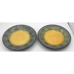 May include: Two ceramic plates with a yellow center and a green rim. The rim features a decorative swirl pattern in a darker shade of green. The plates are round and appear to be handmade.